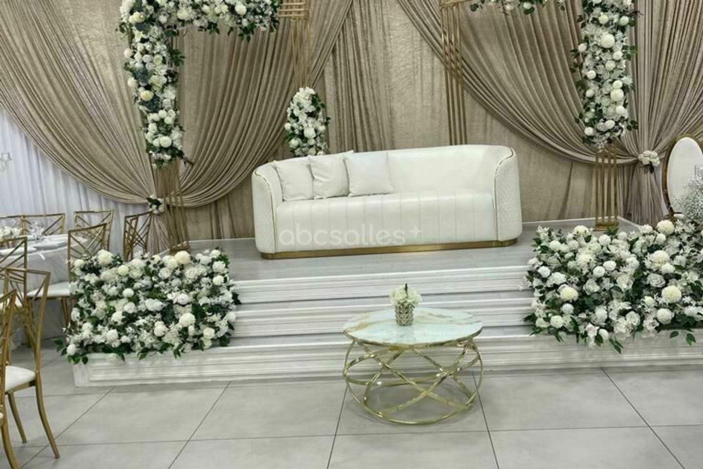 Estrade des mariés : canapé blanc, arches dorées couvertes de roses blanches, voilages champagne.