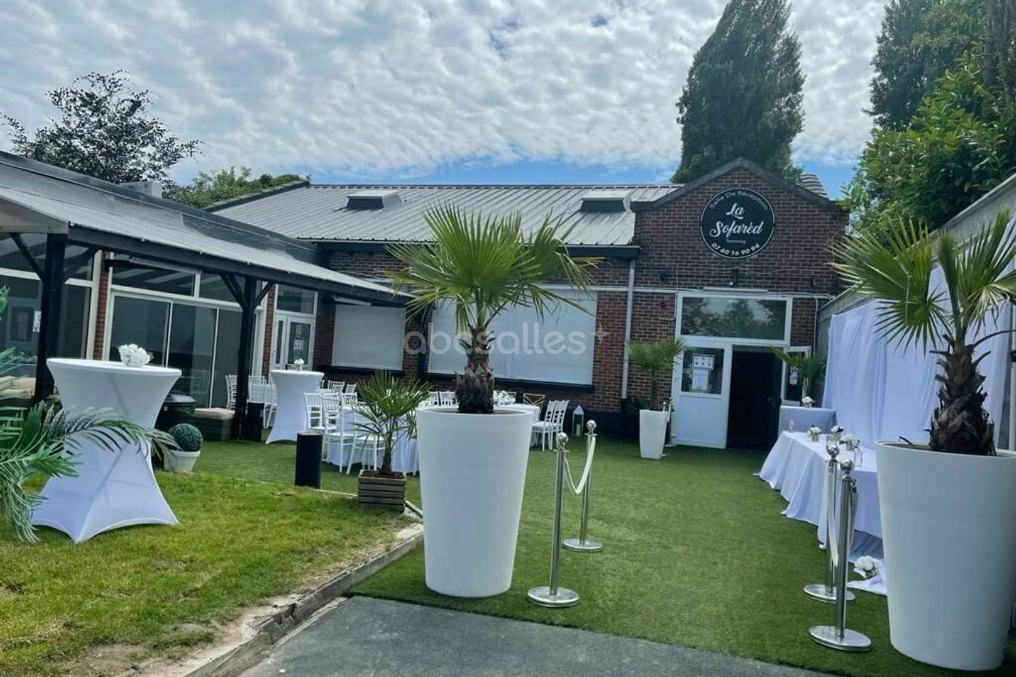 L'entrée de La Sofared un jour de fête : façade de brique, enseigne ronde, palmiers et mange-debout blancs sur la pelouse.